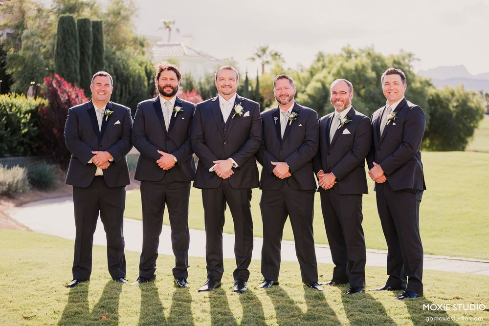 Groomsmen in black suits stand in line on a grassy lawn during an outdoor wedding celebration.
