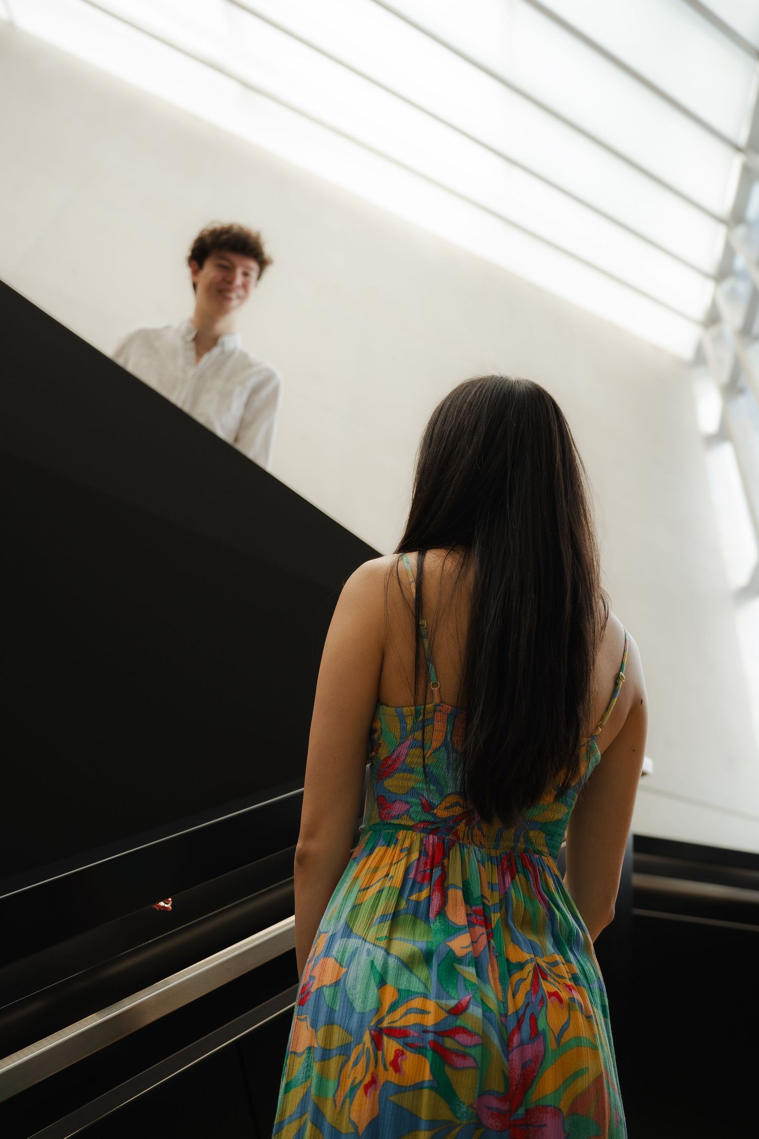 A couple in a colorful summer dress and white shirt pose together in a bright interior setting.