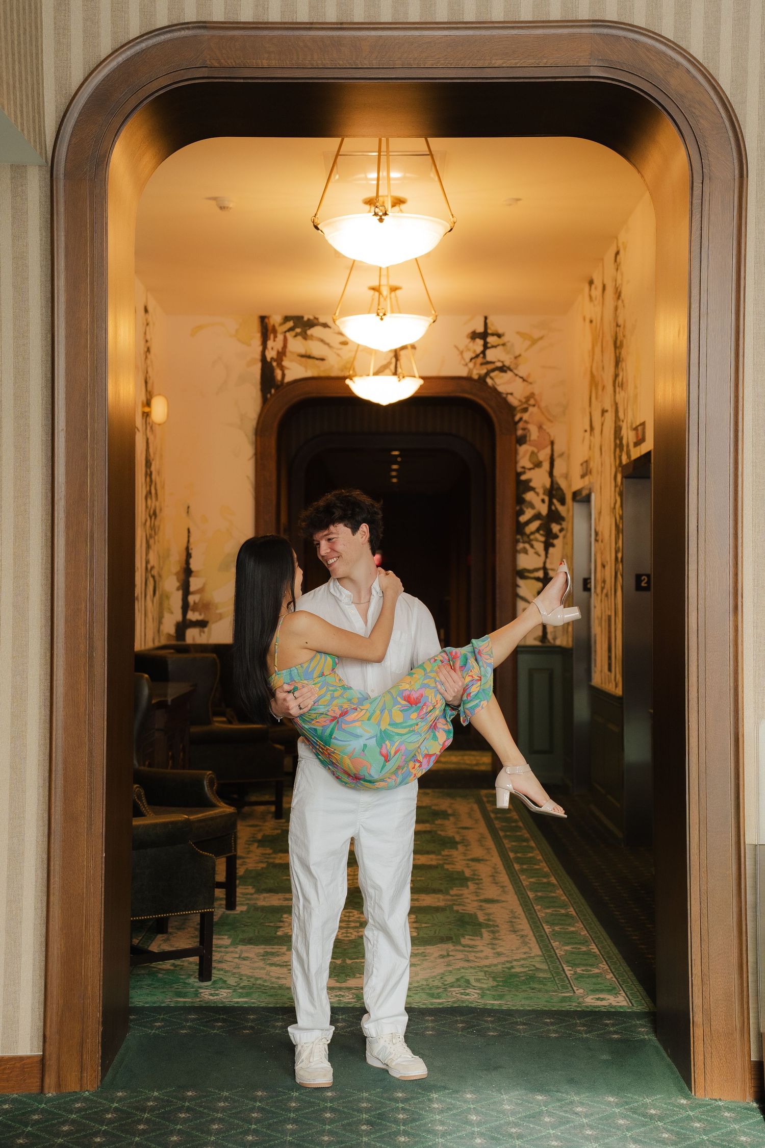 Two people in white and colorful attire dancing playfully in an elegant hallway with chandeliers.