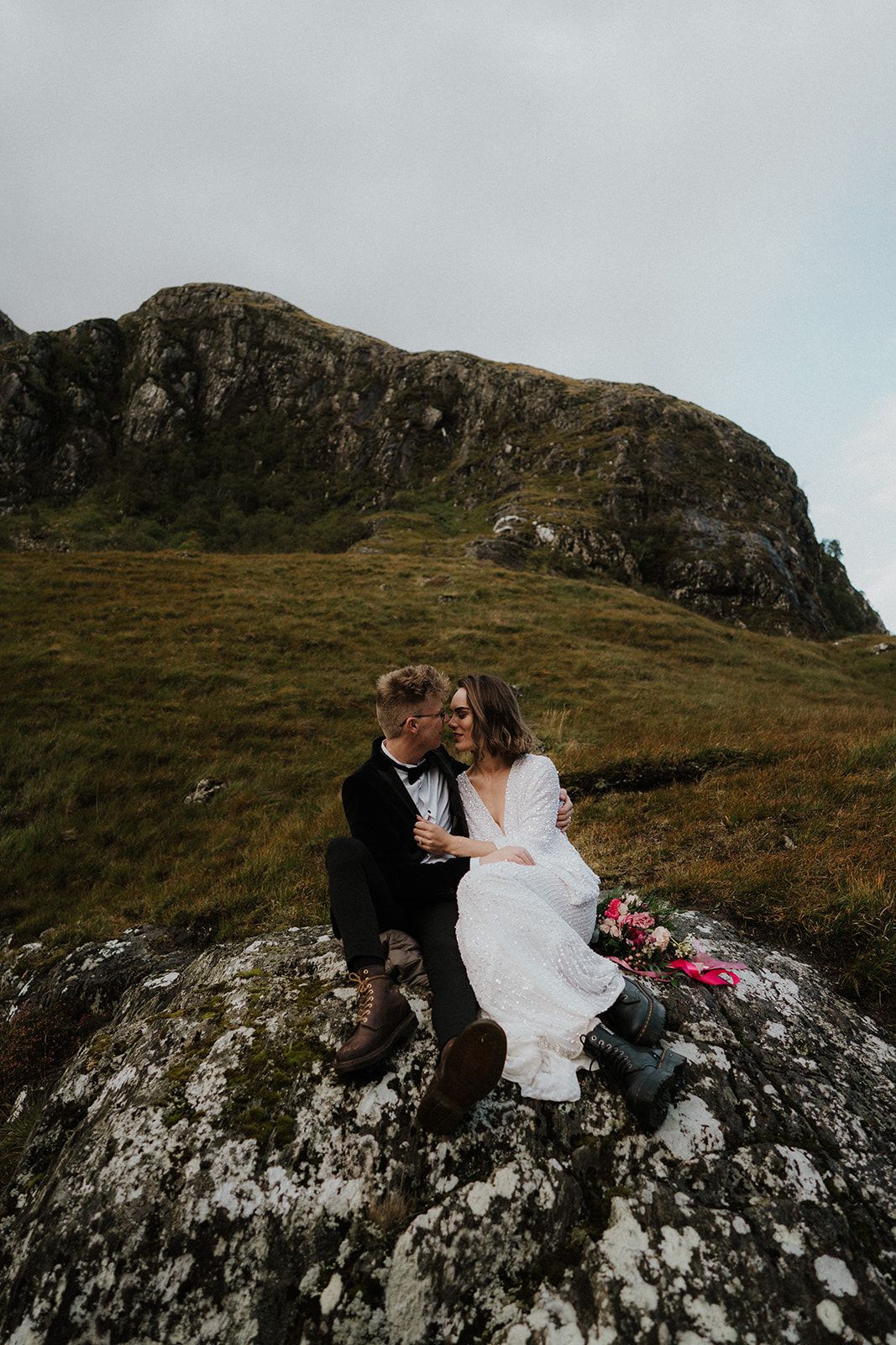 A couple in wedding attire sits intimately on a rocky outcrop against a dramatic Scottish highland landscape.