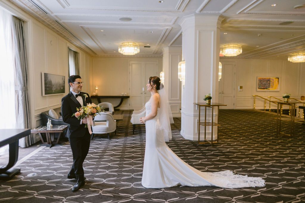 People in formal attire dance in a large carpeted ballroom with ornate white walls and ceiling lights.
