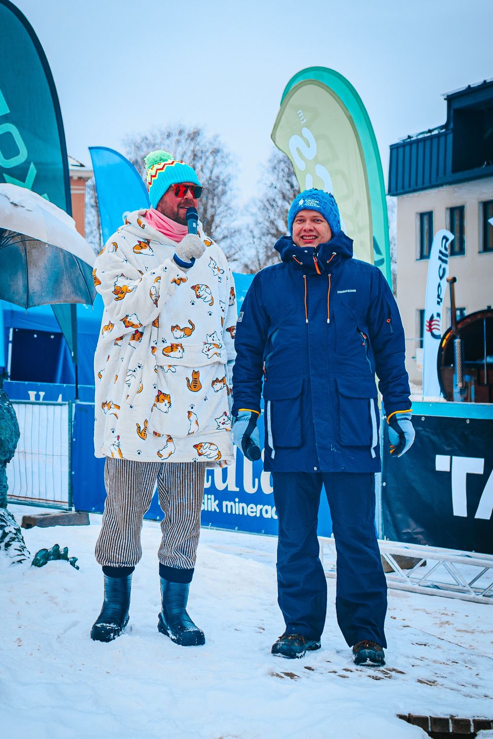 People wearing winter coats and snow gear stand together in snowy conditions at a winter event with blue banners.