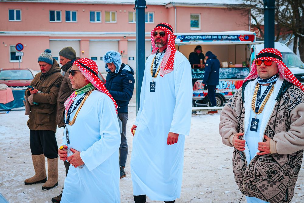 A group in winter coats and red scarves stand in a snowy outdoor area near a pink building.