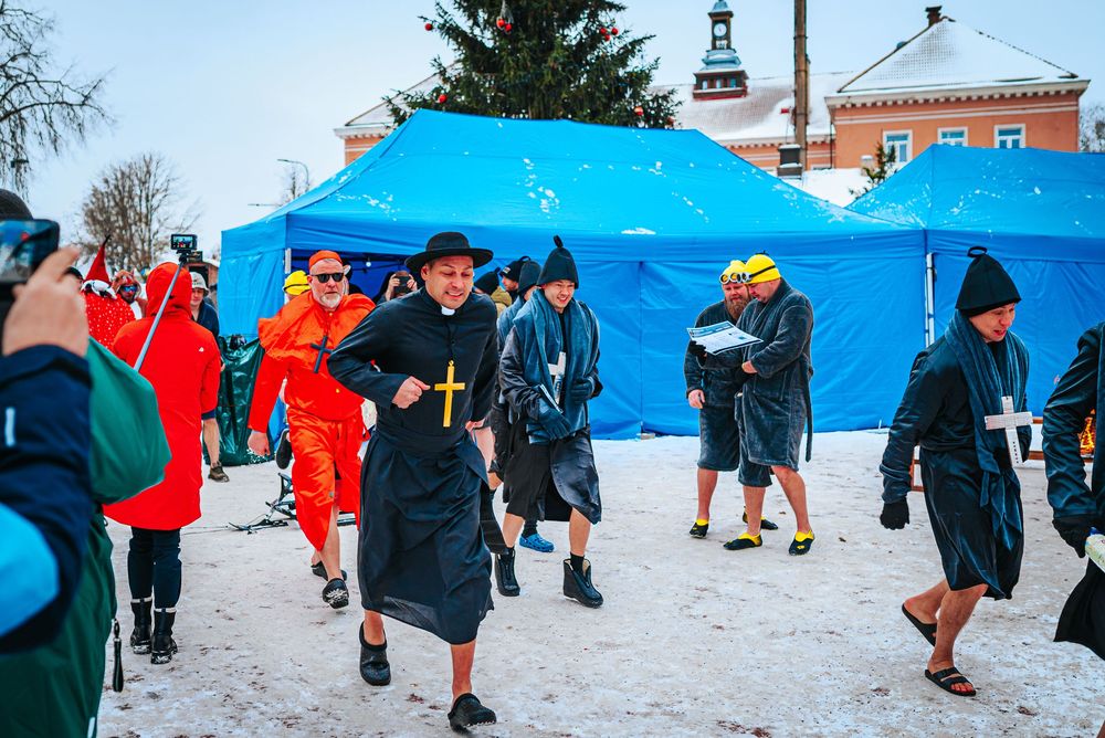Participants in traditional black attire walk past blue tents during a winter festival event in snowy conditions.