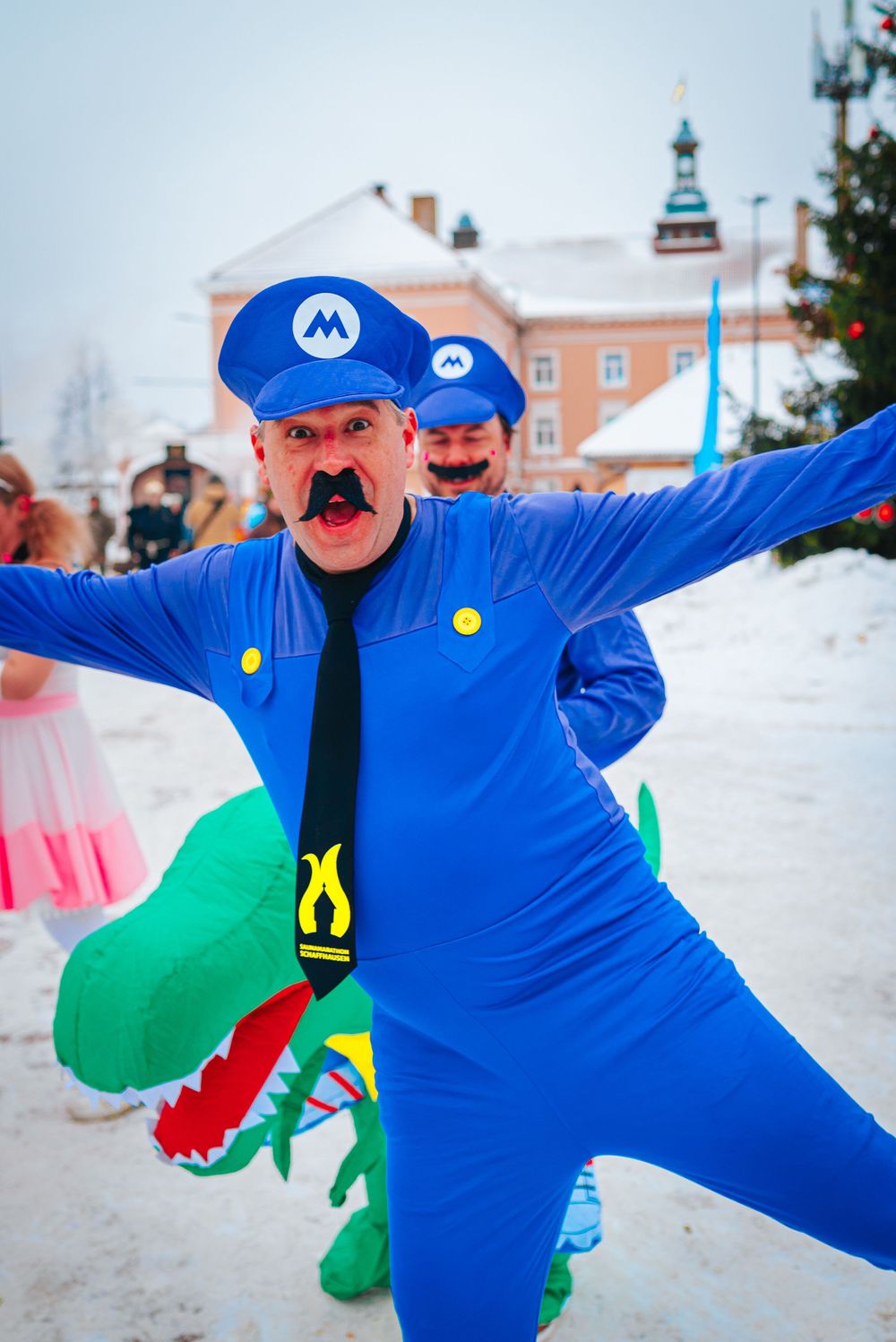 Person in bright blue costume with red mustache performs playful poses in snowy winter setting.