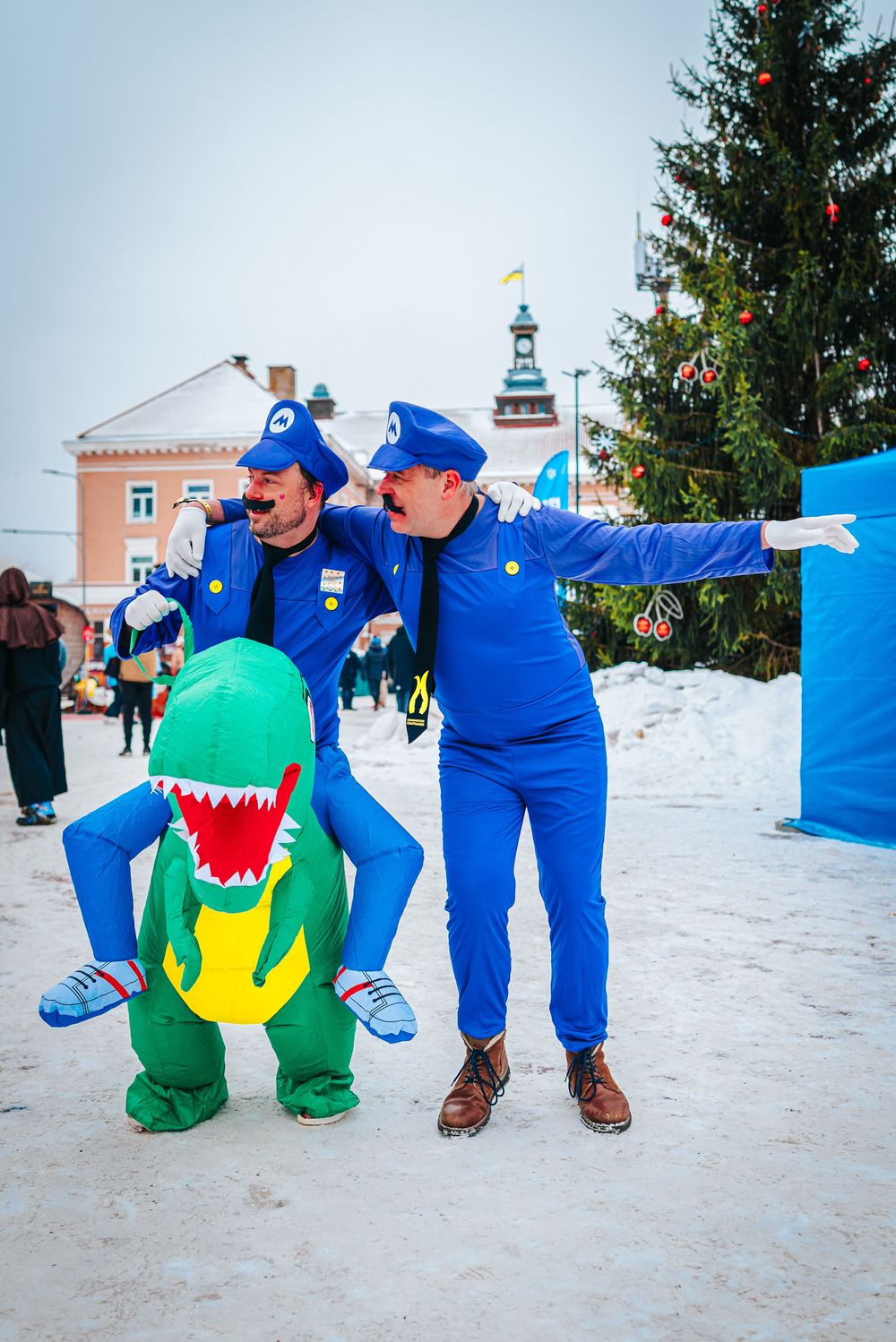 Two performers in bright blue costumes playfully interact with a green dinosaur character in a snowy winter setting.