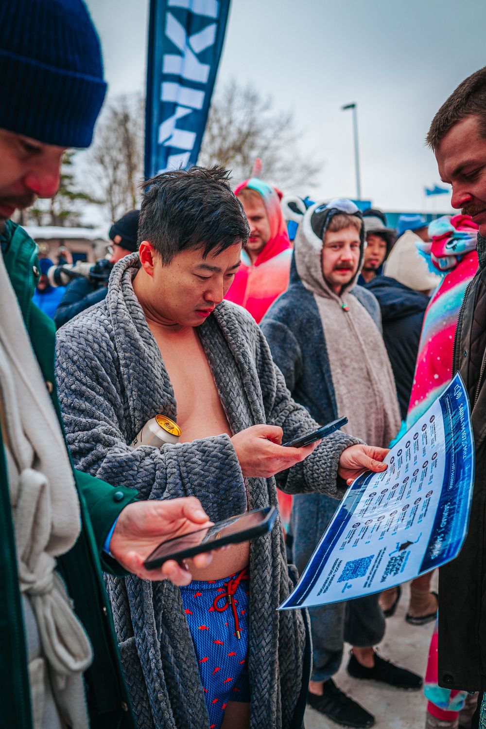 A group of swimmers in winter coats check their phones while gathered outside at a polar plunge event.