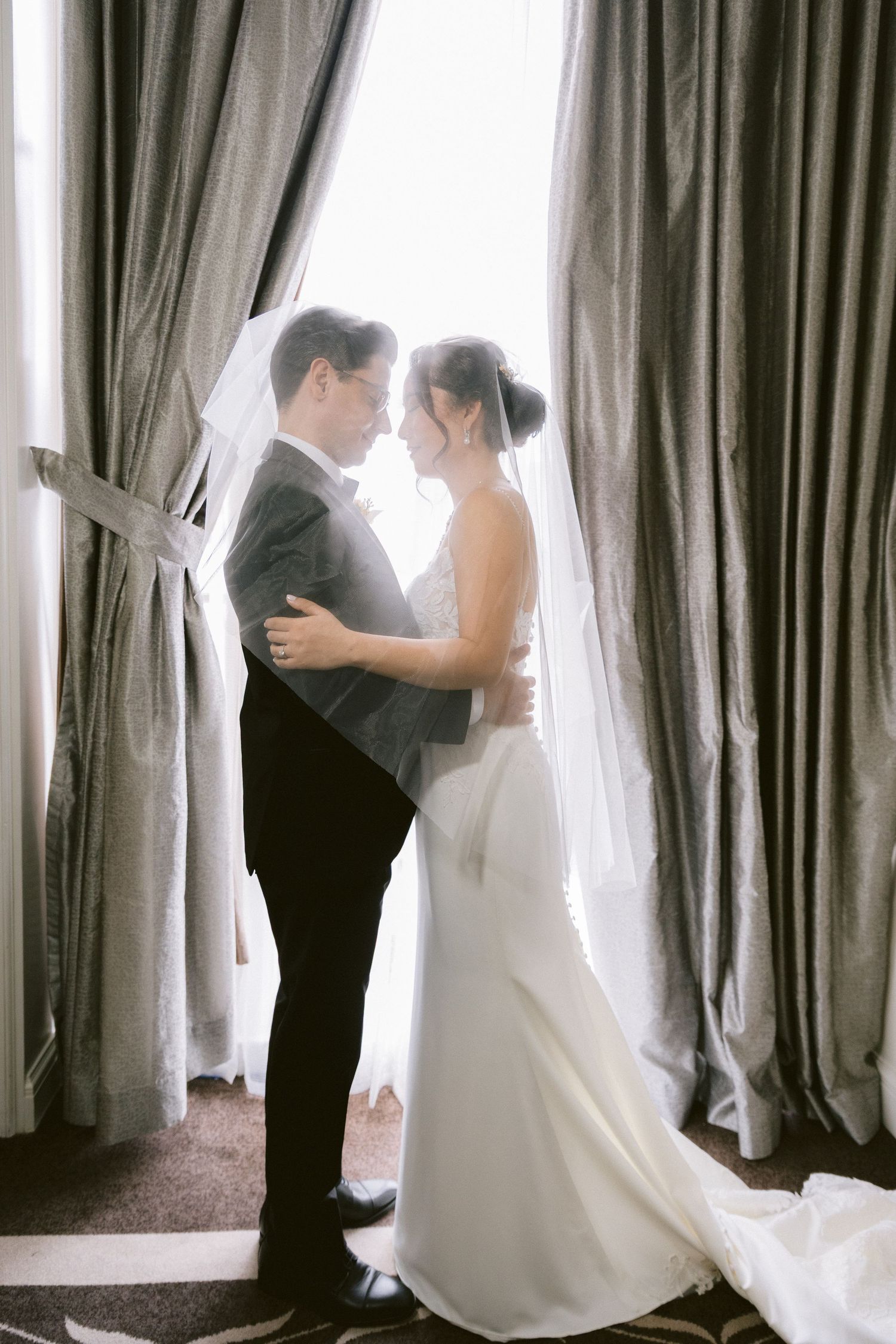 A romantic silhouette of a bride and groom dancing near a window with flowing curtains in a wedding venue.