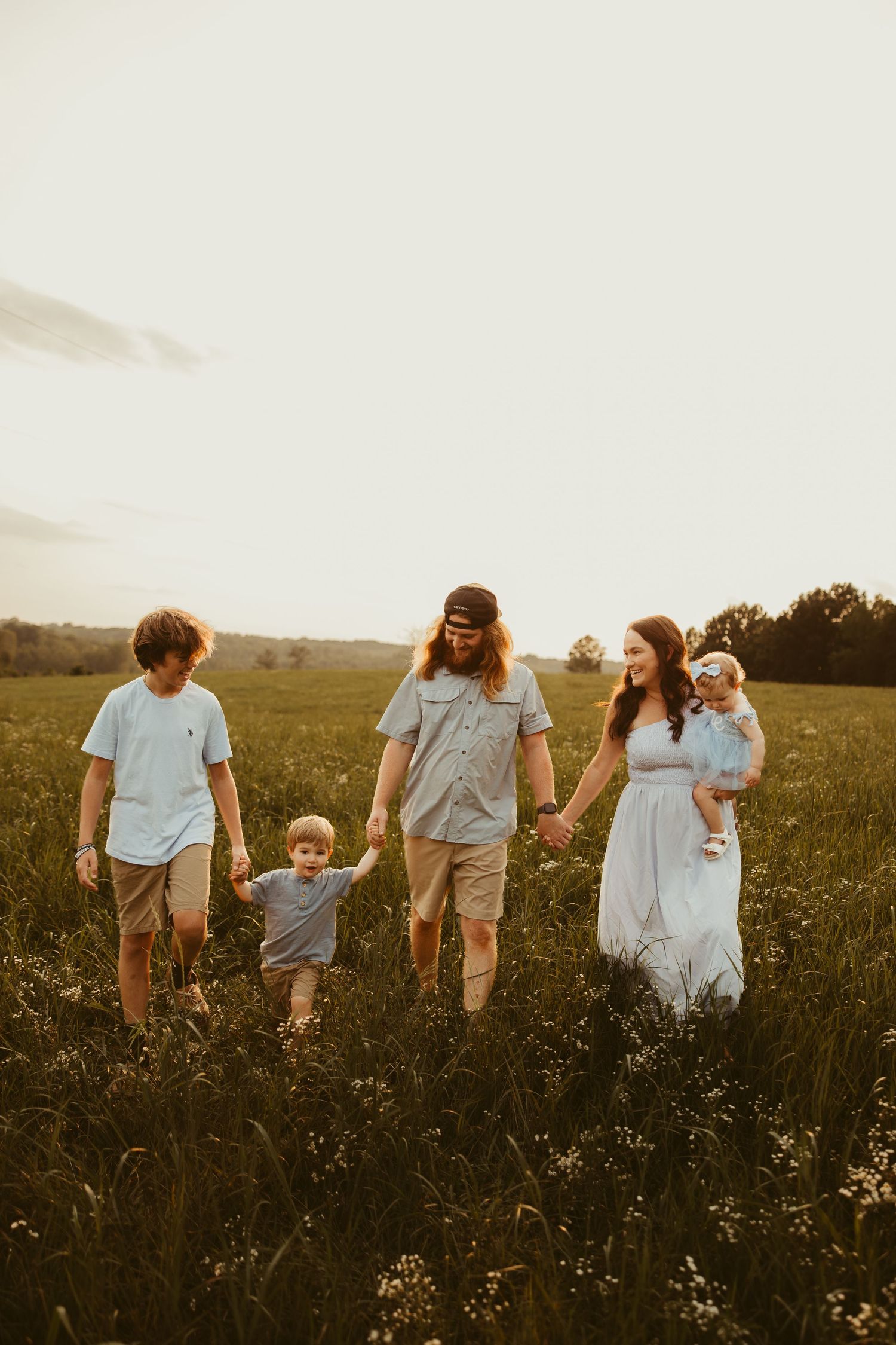 Family of five walking hand-in-hand on a dirt path surrounded by autumn trees and golden hour light. Greensboro Family Photographer