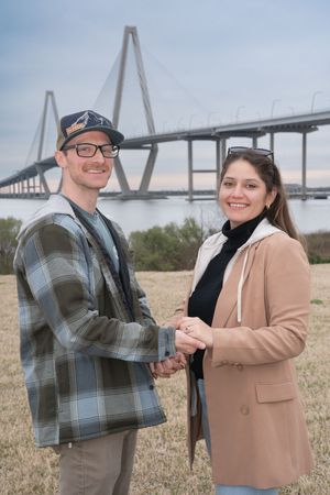 Two people share a happy moment outdoors with a large bridge structure visible in the background.