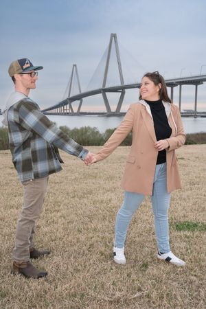 A couple holds hands while standing in a field with a large bridge structure visible in the distance.