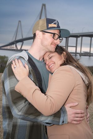A joyful embrace shared between a couple with the Ravenel Bridge visible behind them.