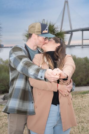 A couple shares a romantic embrace near the iconic Arthur Ravenel Jr. Bridge in Charleston during sunset.