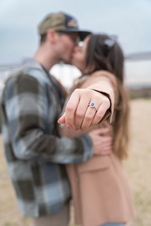 A close-up of an engagement ring while a couple embraces in the blurred background.