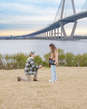 A romantic proposal scene unfolds on a grassy hill with the Arthur Ravenel Jr. Bridge in Charleston as a backdrop.