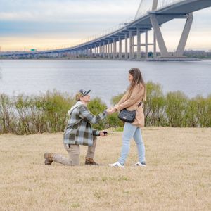 A romantic proposal scene unfolds near a large bridge with grassy waterfront in the background.