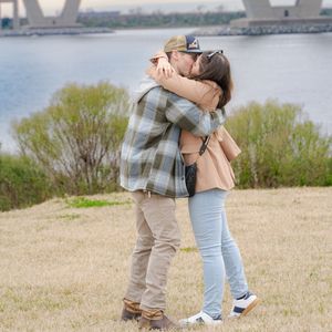 A couple embraces on a grassy hill with a bridge visible in the background.