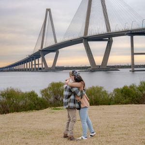 Romantic proposal scene unfolds on hilltop with iconic Ravenel Bridge spanning across Charleston harbor in background.