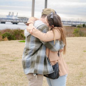 A couple embraces in a tender moment by the waterfront with a bridge visible in the distance.