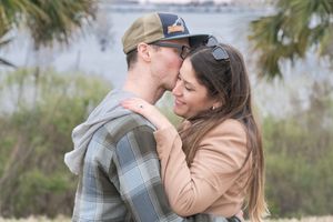 A couple embraces while standing in a grassy field with palm trees and water visible in the background.