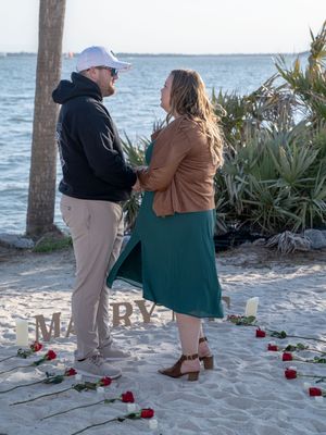 A couple stands together on a sandy beach with red roses scattered at their feet during a romantic moment by the ocean.