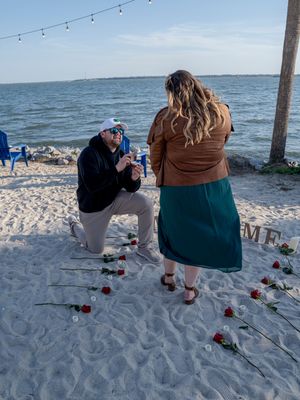 People walking on a sandy beach at sunset with red roses scattered on the ground and string lights overhead.
