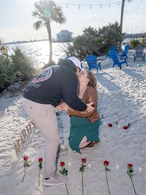 A sequence of photos showing someone proposing on a sandy beach with red roses laid out in a line at sunset.