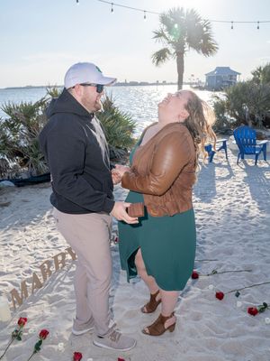 A romantic beach proposal scene unfolds as a couple sits on the sand with string lights and red roses in the background.