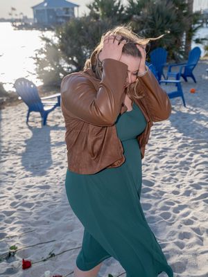 A couple in a green dress and brown jacket embrace romantically on a sandy beach at sunset.
