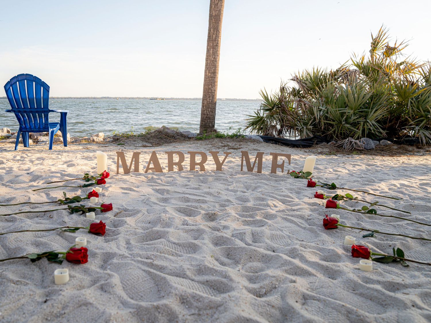 A beach marriage proposal setup with 'MARRY ME' letters in the sand, red roses, and a blue Adirondack chair by the ocean.