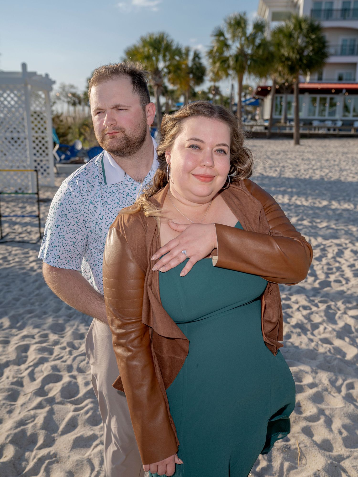 A couple embracing on a sandy beach, one wearing a brown leather jacket and the other in a green dress.