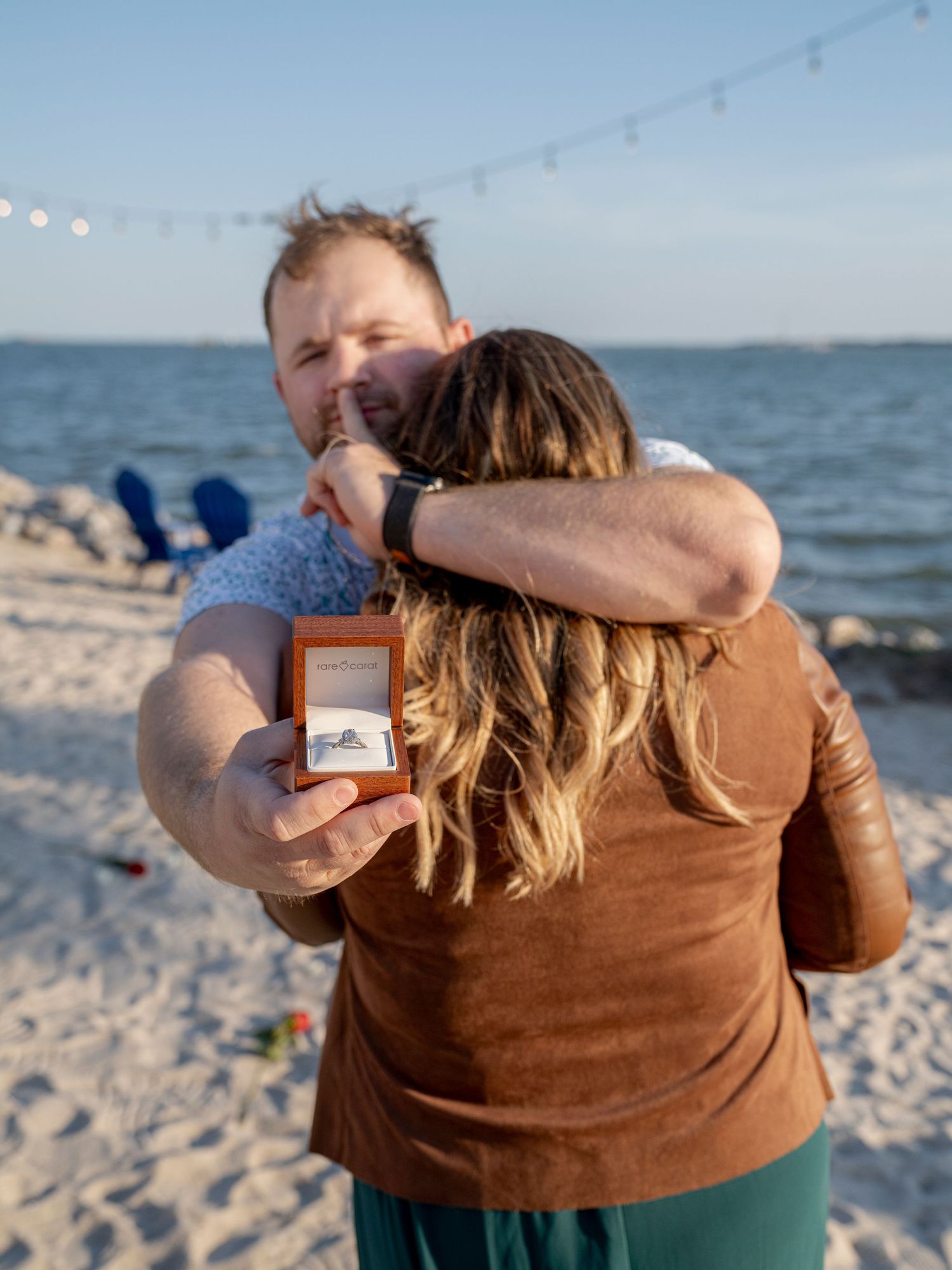 A couple embraces on a sunny beach while sharing a romantic proposal moment with an engagement ring.