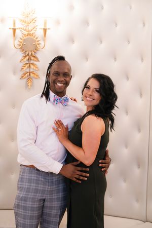 A couple embraces and smiles in front of an elegant white tufted wall with gold sconces.