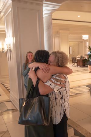 Two people share warm hugs in a bright hotel lobby with white columns and marble floors.