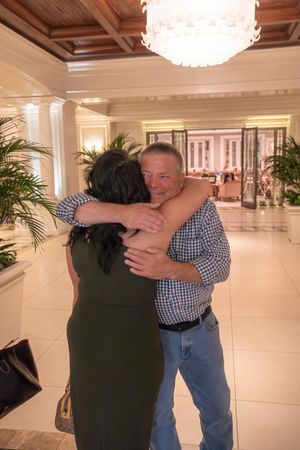 Two people embrace in an emotional reunion inside a bright, spacious building hallway with white walls and floors.