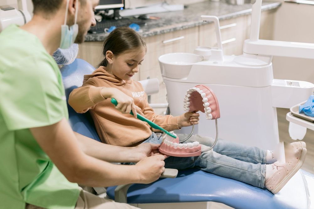 A young girl in a dental chair learning to brush teeth on a model jaw with a dentist in green scrubs.