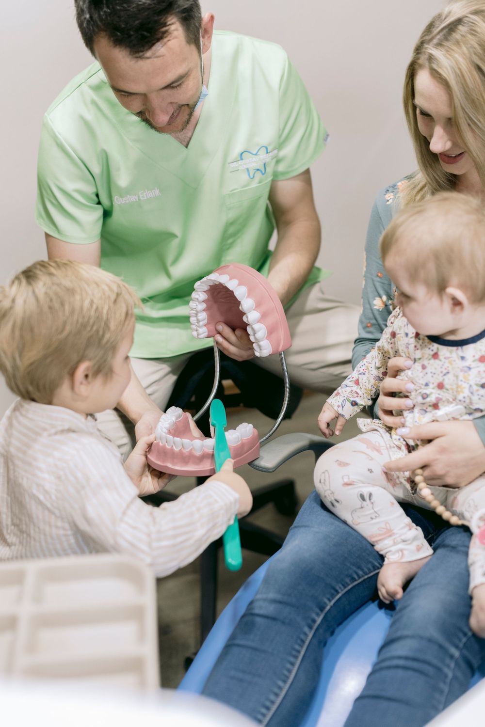 A dentist in green scrubs showing children a dental jaw model while a mother watches in a dental office.
