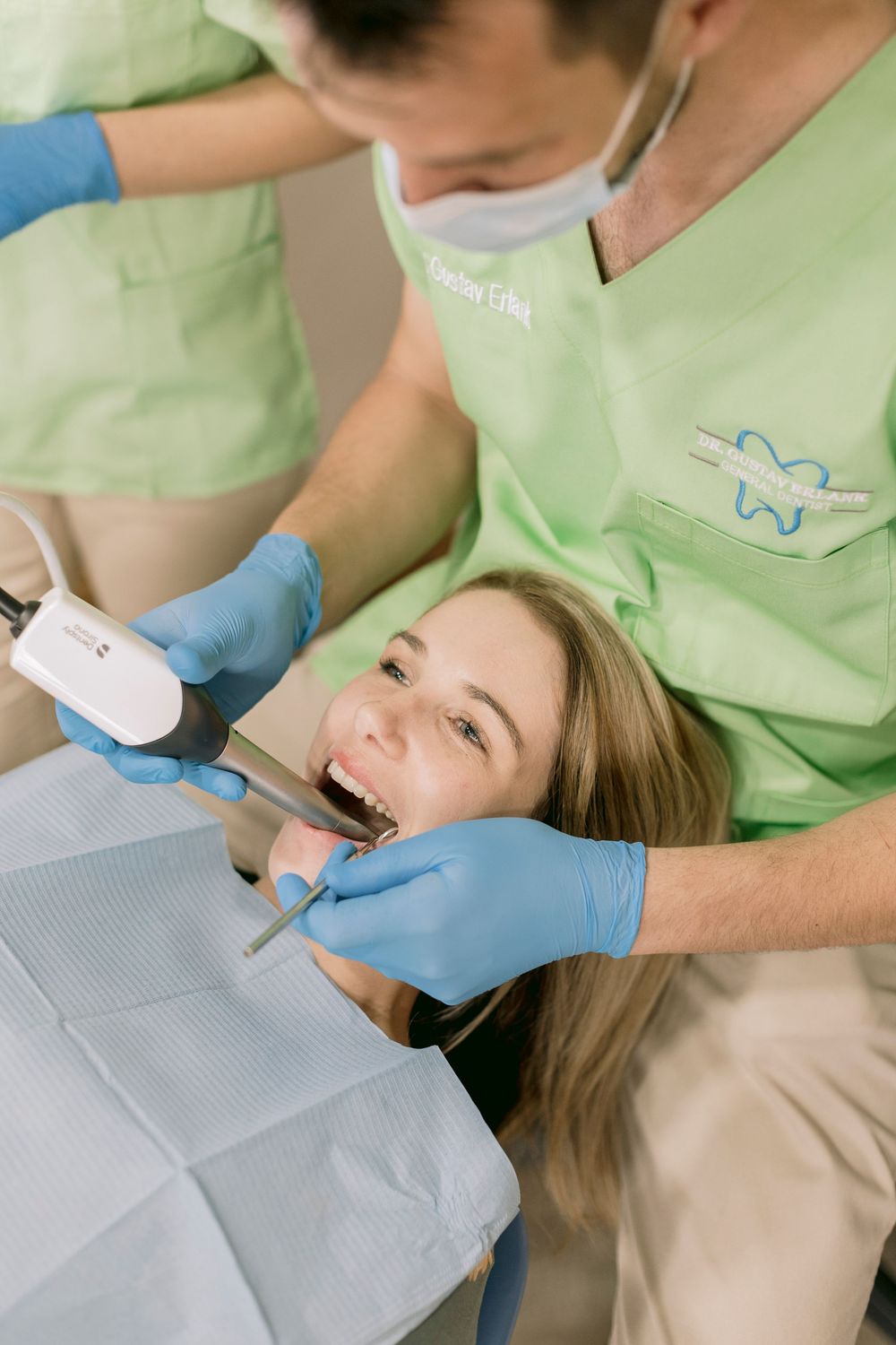 Dentist in green scrubs and blue gloves performing dental examination on smiling female patient.
