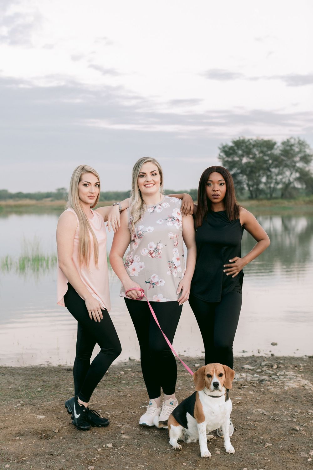 Three diverse women in athletic wear posing outdoors near a lake with a beagle on a pink leash.