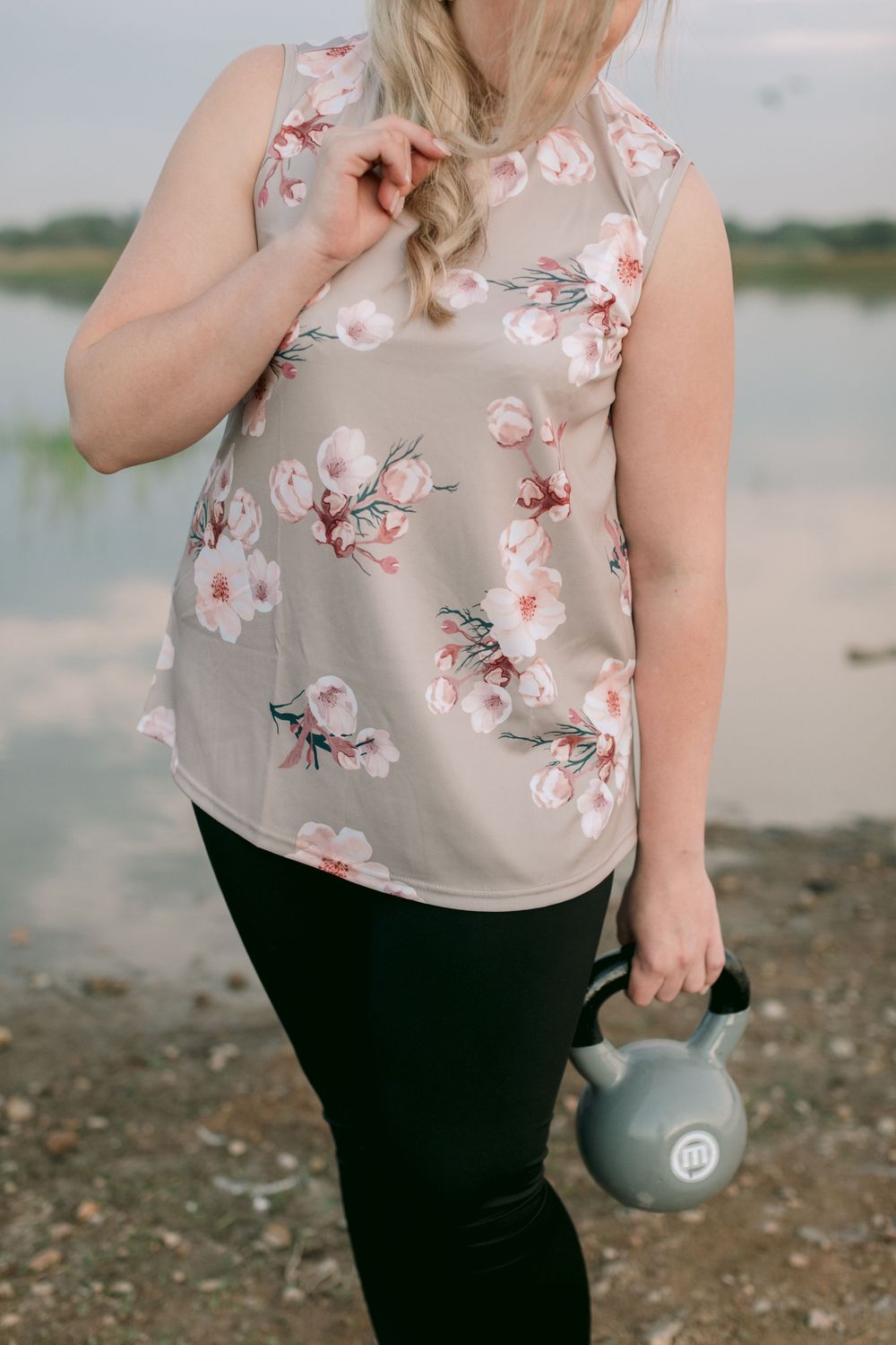 Woman in floral sleeveless top holding a kettlebell while standing near a reflective lake outdoors.