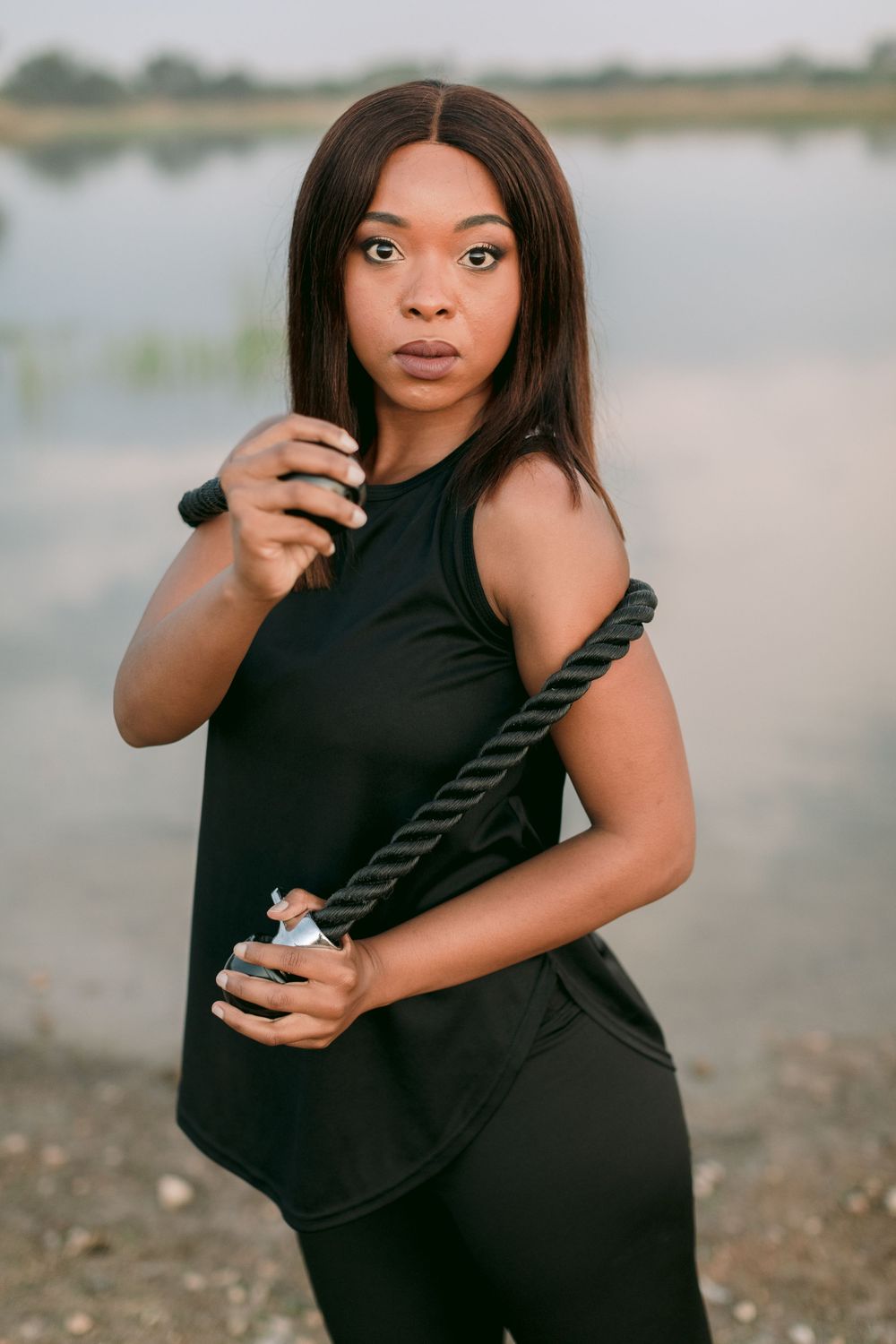 Confident Black woman in black athletic wear posing with a battle rope near a serene lake.