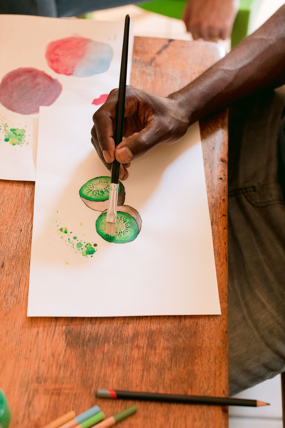Close-up of a person painting watercolor kiwi fruit slices on white paper.