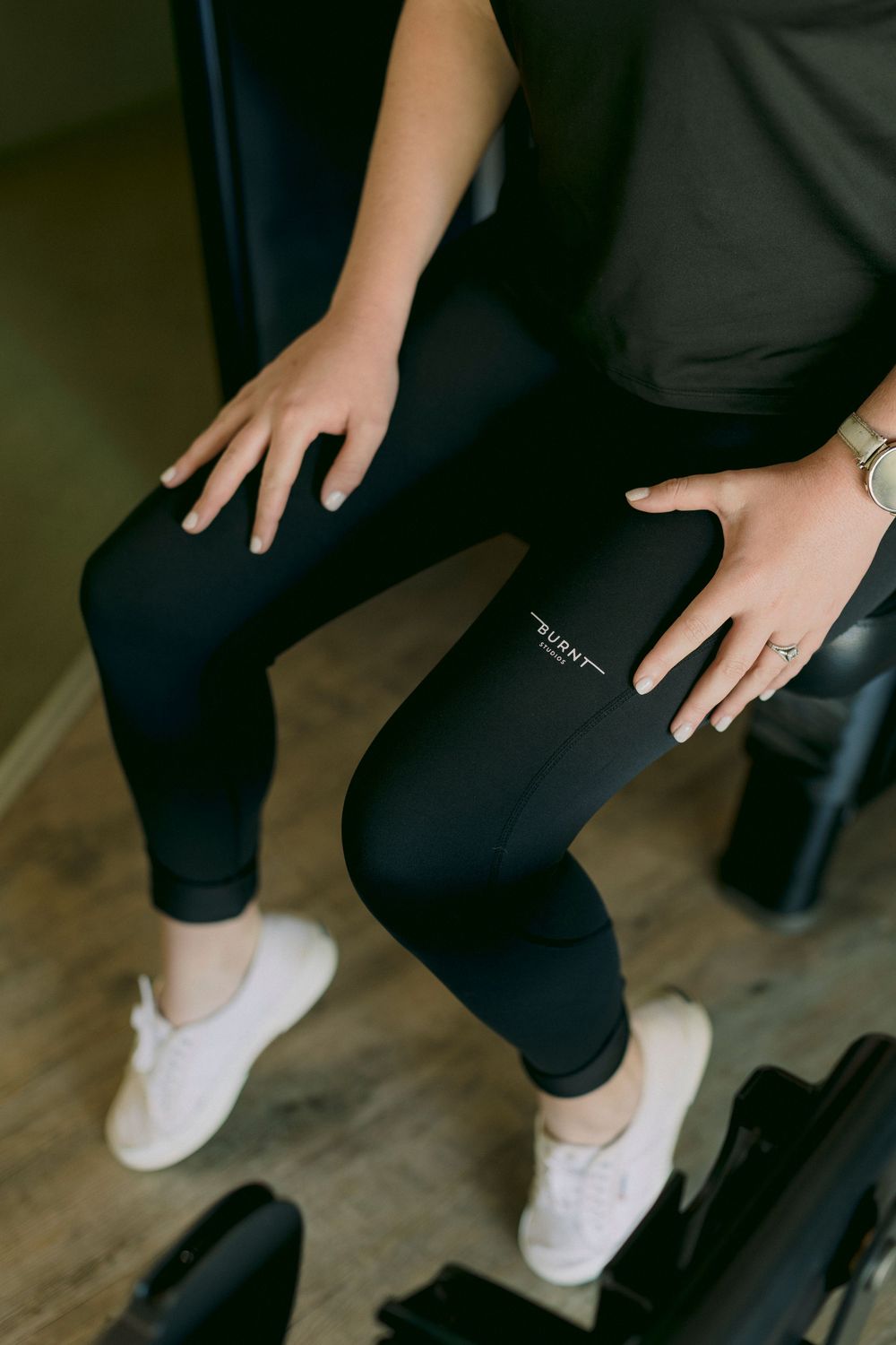 Close-up of a woman wearing black leggings and white sneakers sitting on gym equipment.