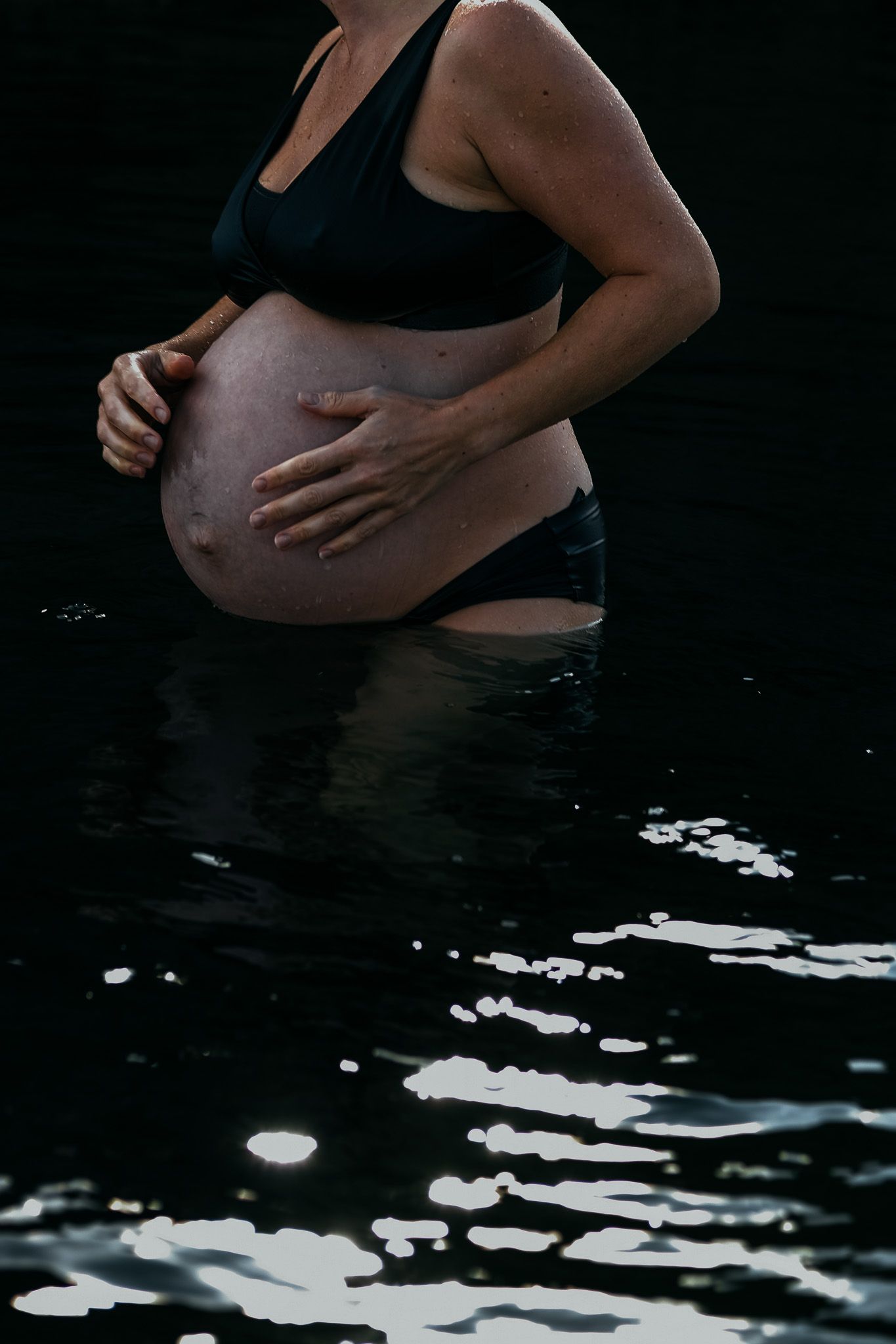 Maternity portrait of an expectant mother standing in water, gently holding her bump during a calm, natural pregnan