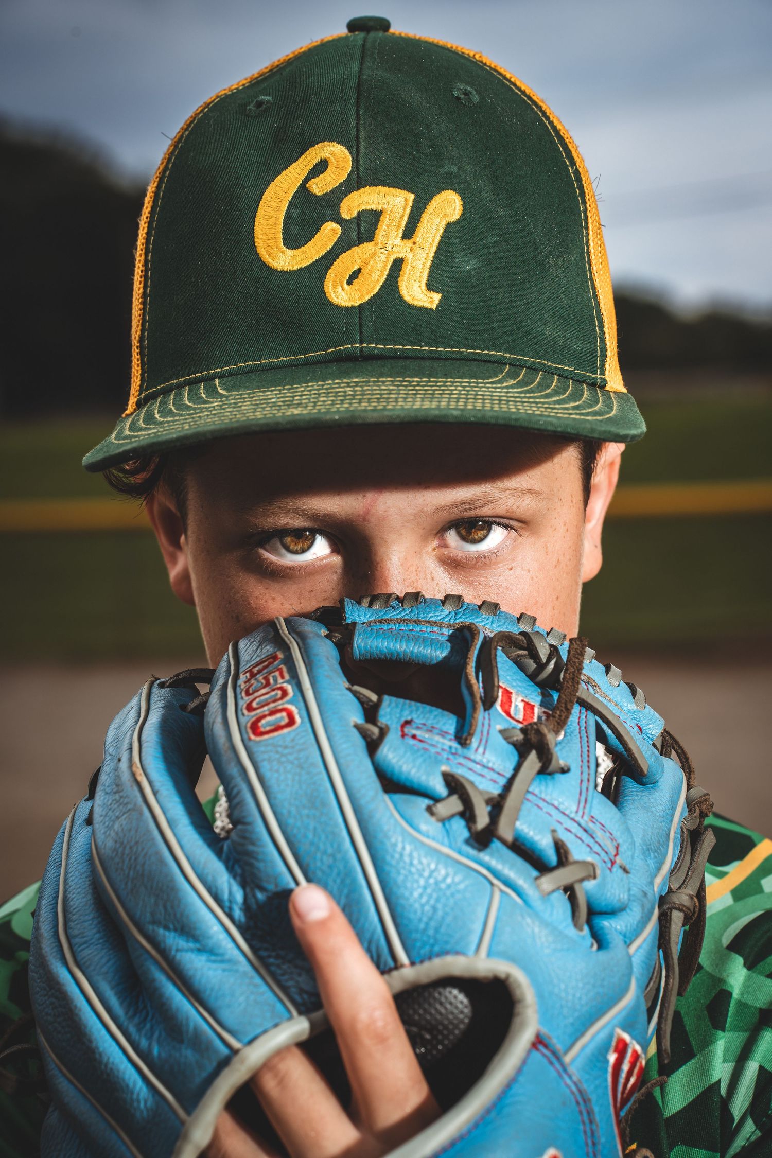 Baseball player in green and yellow cap peers over light blue baseball glove in dramatic sports portrait.