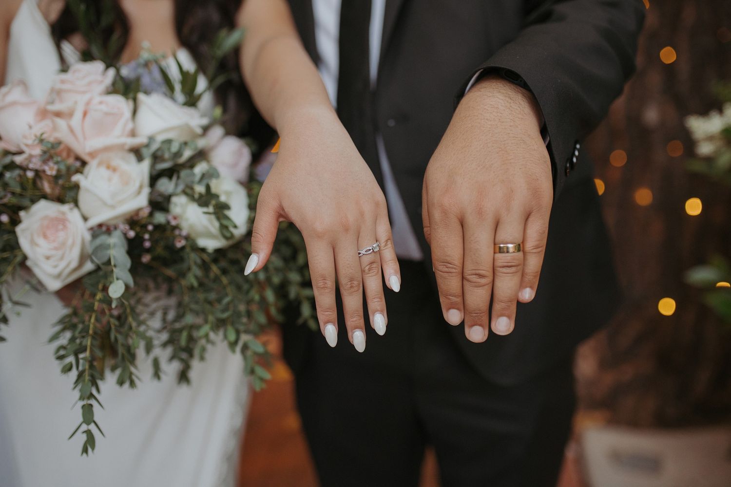 Close-up view of hands displaying wedding rings with elegant floral bouquet in soft pink and white tones in the background.