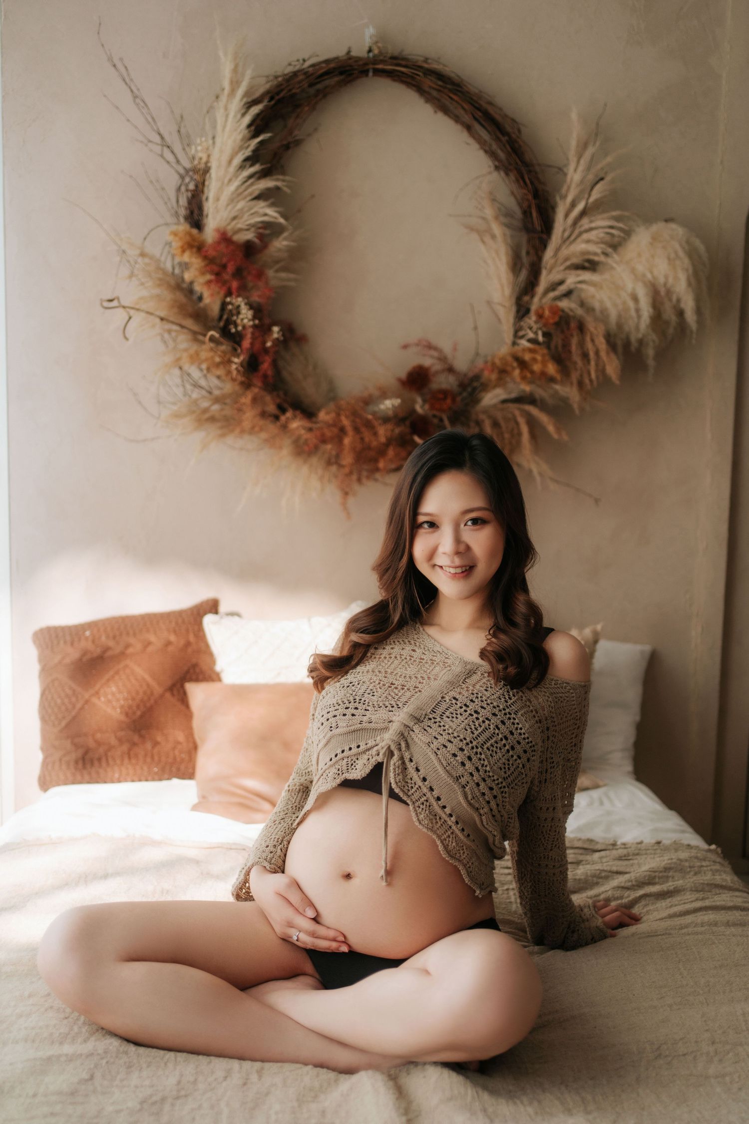 Maternity photo in warm tones featuring a knit sweater and dried floral wreath wall decor in a cozy bedroom setting.