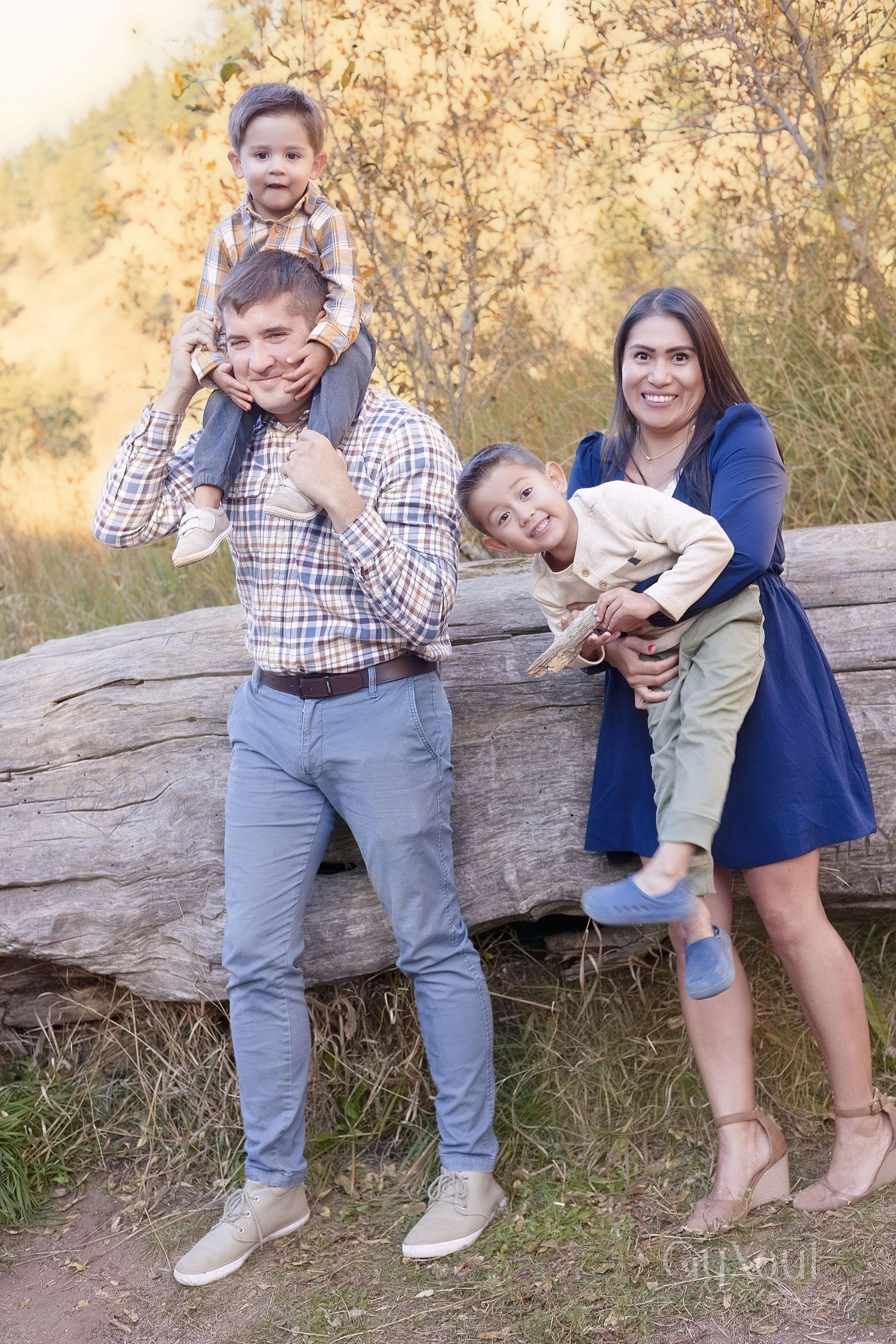 A family poses together outdoors on a fall day near a large rock with golden foliage in the background.