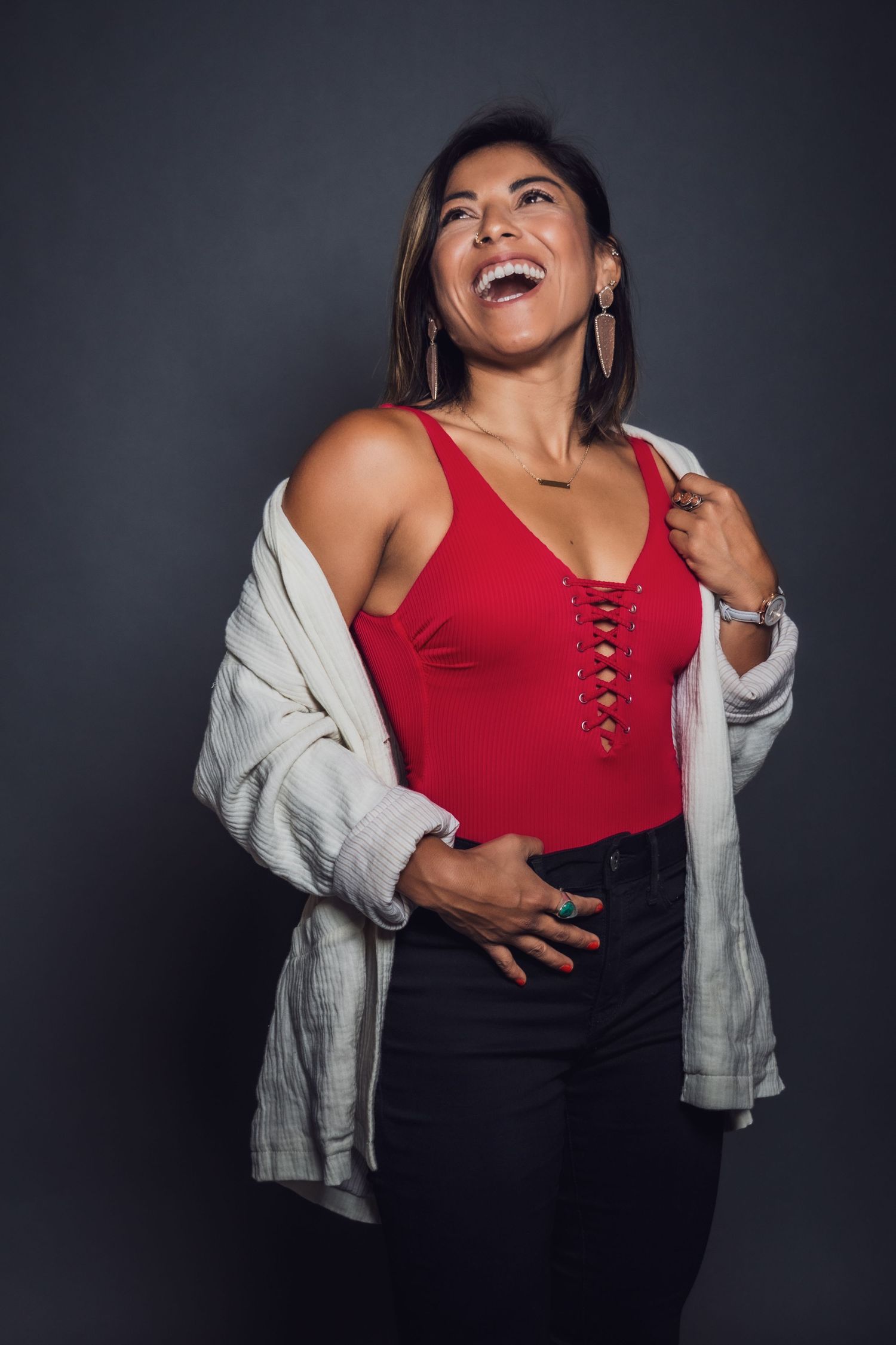 Laughing woman in red lace-up top, white cardigan, and black jeans posing against a dark gray background.
