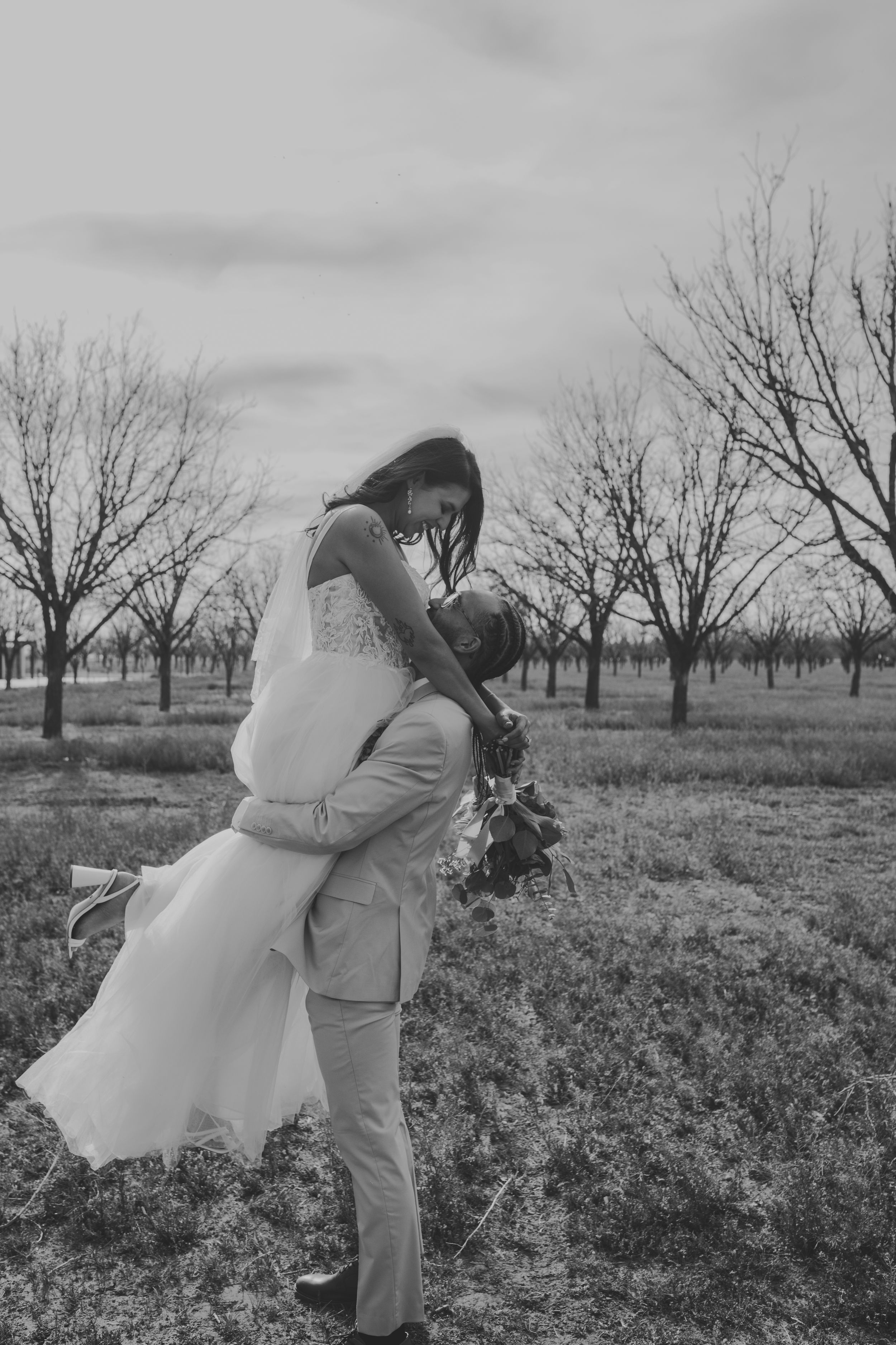 Black and white photo of groom lifting bride in a field of bare trees on their wedding day.